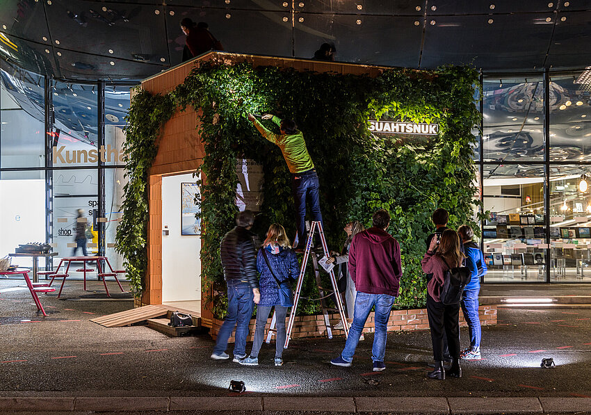 Menschen versammeln sich vor dem Kunsthaus Graz in der Nacht. Am Vorplatz steht ein kleines Gebäude, das mit Hopfen-Pflanzen überwachsen ist. Der Künstler Alfredo Barsuglia steht auf einer Leiter und erntet den Hopfen.