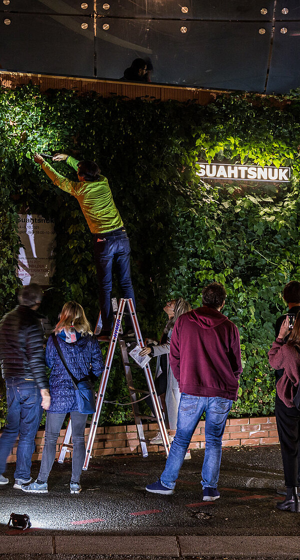 Menschen versammeln sich vor dem Kunsthaus Graz in der Nacht. Am Vorplatz steht ein kleines Gebäude, das mit Hopfen-Pflanzen überwachsen ist. Der Künstler Alfredo Barsuglia steht auf einer Leiter und erntet den Hopfen.