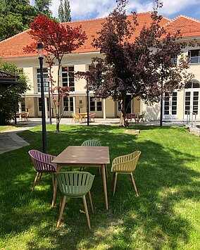 Blick Richtung Heimatsaal im Duftgarten. Im Vordergrund ist ein Tisch mit Sesseln, seitlich sieht man den Pavillon.