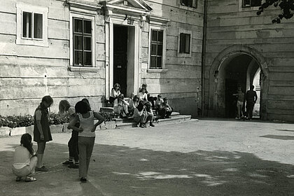 Eine schwarz-weiß Fotografie von Jugendlichen, auf einem Hof. Eine Gruppe steht links im Vordergrund, eine andere sitzt auf einer Treppe weiterhinten.