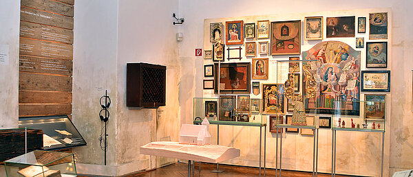 The photo shows an exhibition room. In the center is a small platform made of bricks, on it is a small model of a chapel, made of milky glass. On the far wall hang numerous pictures (paintings, votives). In front of them are three showcases with objects from the religious context, for example, a statue of the Virgin Mary and votive offerings made of red wax.