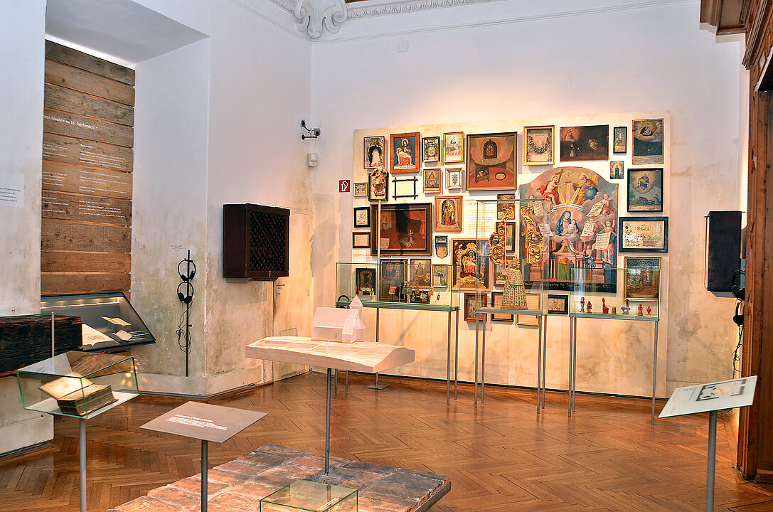 The photo shows an exhibition room. In the center is a small platform made of bricks, on it is a small model of a chapel, made of milky glass. On the far wall hang numerous pictures (paintings, votives). In front of them are three showcases with objects from the religious context, for example, a statue of the Virgin Mary and votive offerings made of red wax.