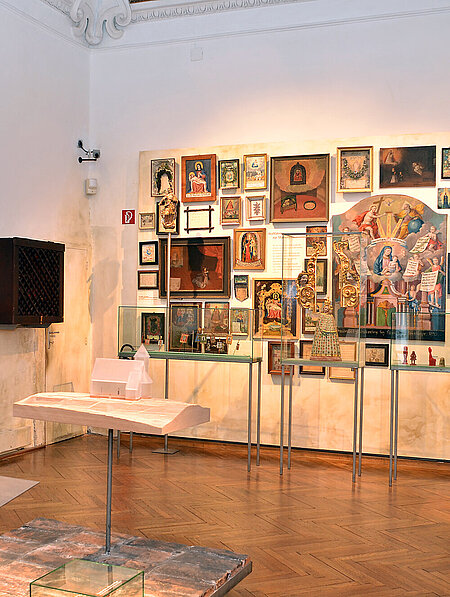 The photo shows an exhibition room. In the center is a small platform made of bricks, on it is a small model of a chapel, made of milky glass. On the far wall hang numerous pictures (paintings, votives). In front of them are three showcases with objects from the religious context, for example, a statue of the Virgin Mary and votive offerings made of red wax.