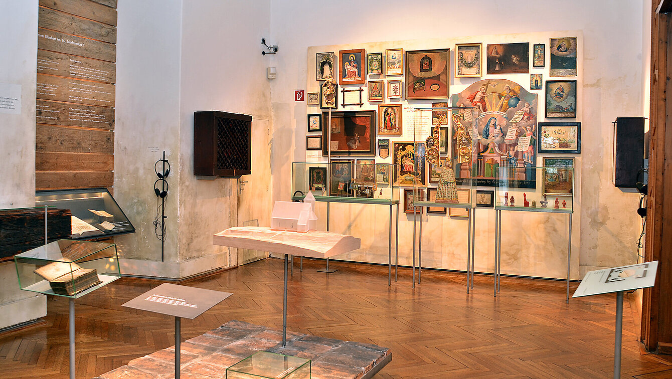 The photo shows an exhibition room. In the center is a small platform made of bricks, on it is a small model of a chapel, made of milky glass. On the far wall hang numerous pictures (paintings, votives). In front of them are three showcases with objects from the religious context, for example, a statue of the Virgin Mary and votive offerings made of red wax.