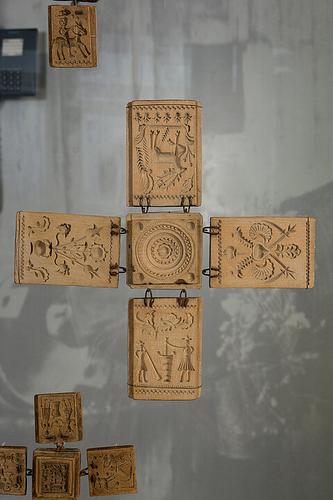 Five rectangular pieces of wood, four of which are attached to the fifth square with their short sides using metal eyelets. The wooden pieces are decorated with embedded people and plants.