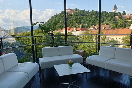 Blick auf den Schlossberg aus der Needle im Kunsthaus Graz. Im Vordergrund stehen stehen weiße Sitzcouchen und ein kleiner Tisch.