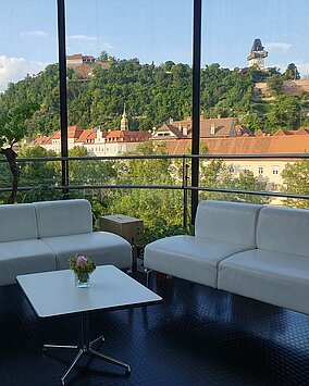 Blick auf den Schlossberg aus der Needle im Kunsthaus Graz. Im Vordergrund stehen stehen weiße Sitzcouchen und ein kleiner Tisch.