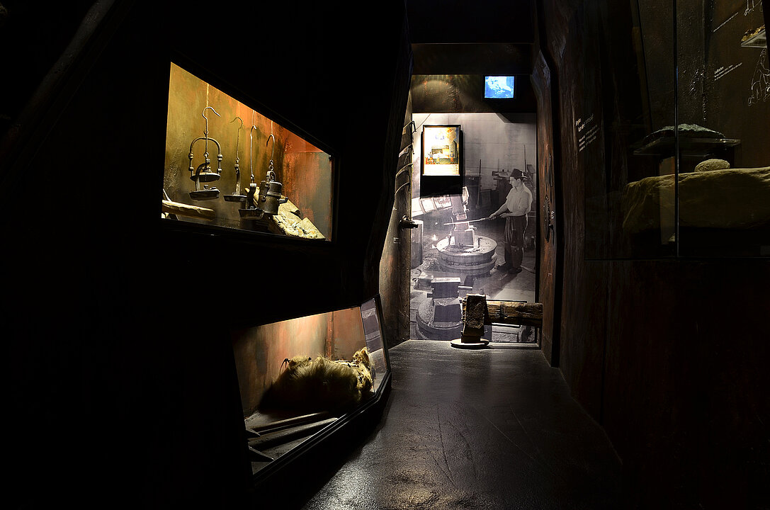 Exhibition view of a dark corridor. To the left and right of the corridor, illuminated showcases are set into the walls, in which miners' work tools can be seen. The corridor is reminiscent of an underground shaft.