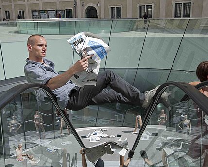 Ein Mann mit Zeitung in der Hand sitzt auf dem Handlauf der Rolltreppe im Joanneumsviertel im Rahmen der Eröffnung der 21. Int. Bühnenwerkstatt.