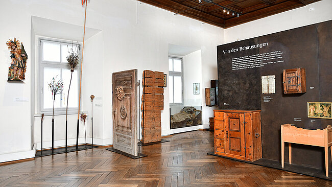 Fotografie von einem Ausstellungsraum. Vor einer weißen und einer braunen Wand stehen verschiedene Gegenstände z.B. Links eine Statue des heiligen Florian, verschiedene Palmbuschen, eine Haustüre mit „Sonnwendkranzerl“