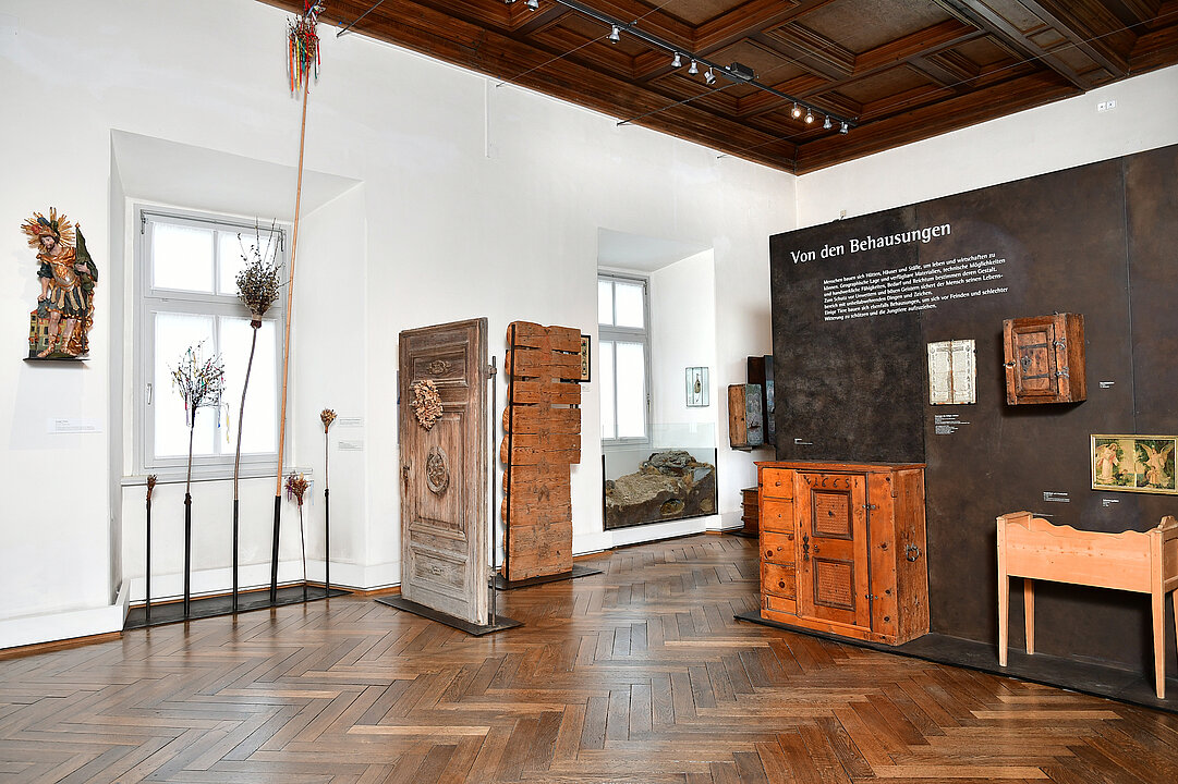 Photography of an exhibition room. In front of a white and a brown wall are various objects e.g. on the left a statue of St. Florian, various palm bouquets, a house door.
