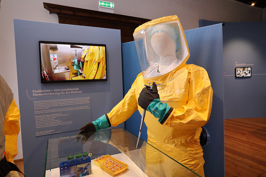  A mannequin with a yellow protective coat stands in front of a blue wall and a screen. The mannequin has a pipette in its hand.