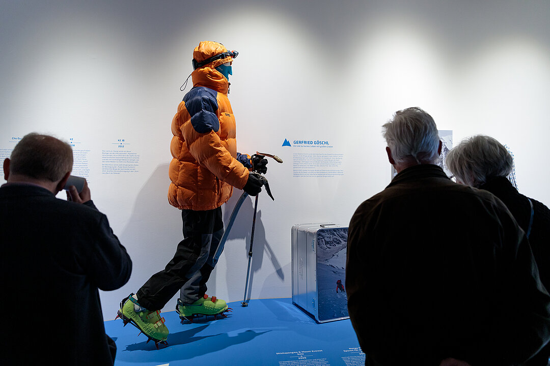 Foto von der Ausstellungseröffnung. Menschen stehen in einem Raum und betrachten eine Schaufensterpuppe im Bergsteiger-Outfit.