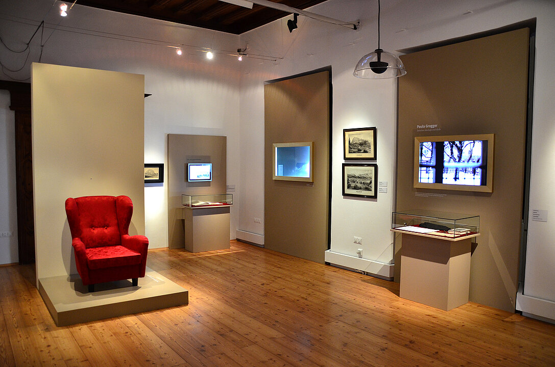 Ausstellungsansicht von einem Raum. Auf einem Sockel steht links im Raum ein roter Sessel.