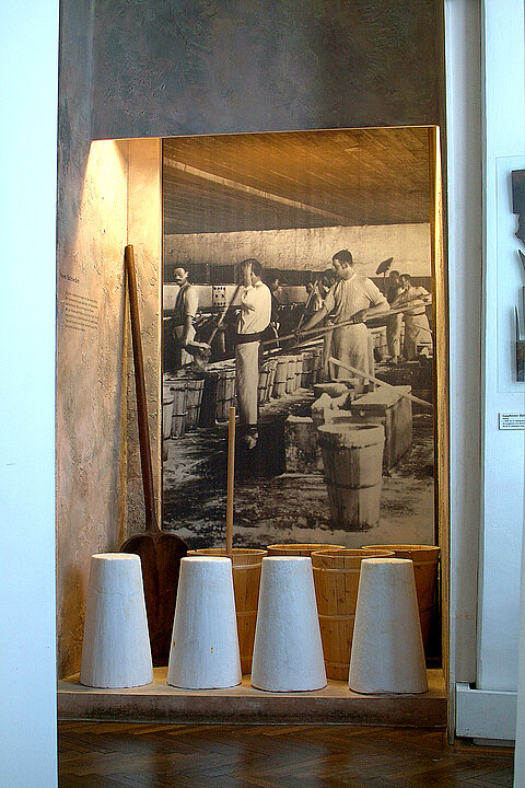 Fotografie von einem Ausstellungsraum. Am Boden stehen vier weiße zylinderförmige Blöcke aus Salz. Dahinter ist eine großflächige Fotografie von Männern, die diese Salzblöcke herstellen.