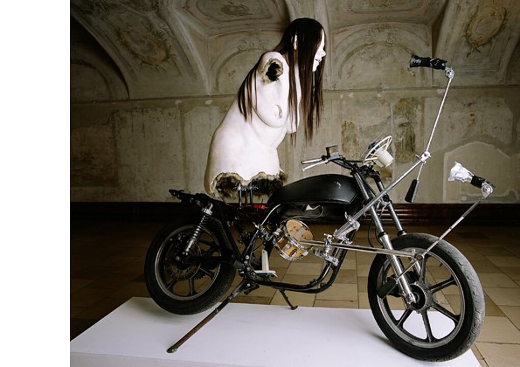 Kunstinstallation: Der Oberkörper einer nackten Frau mit langen schwarzen Haaren sitzt auf einem schwarzen Motorrad.