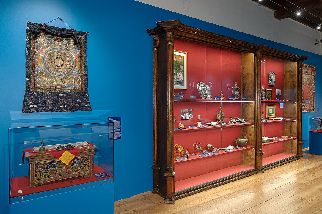 Ausstellungsansicht von einer blauen Wand, vor der eine hohe Vitrine aus Holz steht. In der Vitrine sind viele kleine Gegenstände. Daneben hängt ein Wandteppich.