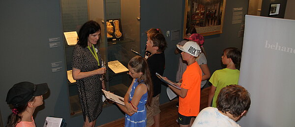 A woman stands in an exhibition room and talks to a group of children.