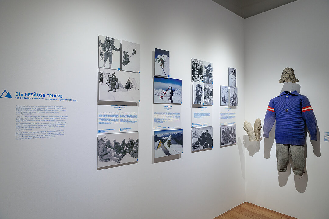 Ausstellungsansicht: An einer Wand hängen Fotografien und ein Outfit mit blauem Pullover, kurzer grauer Hose und einem Hut.