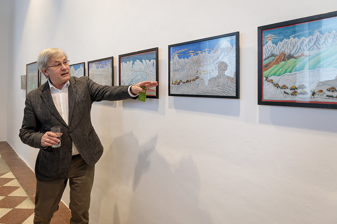 Ein Mann zeigt mit einer Hand auf verschiedene gerahmte Zeichnungen, die vor einer weißen Wand hängen. 

