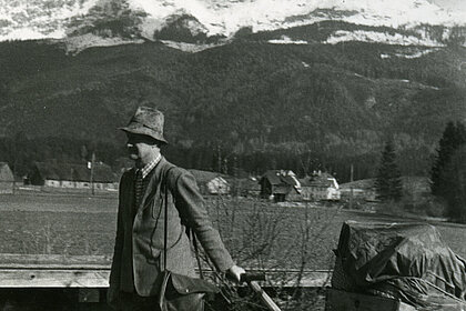 Ein Mann mit Hut zieht einen Handwagen, auf dem umwickelte Gegenstände liegen. Im Hintergrund sieht man Berge und Wälder.