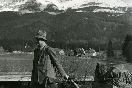 A man pulls a handcart with wrapped objects lying on it. In the background you can see mountains and forests.