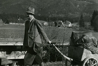 A man pulls a handcart with wrapped objects lying on it. In the background you can see mountains and forests.