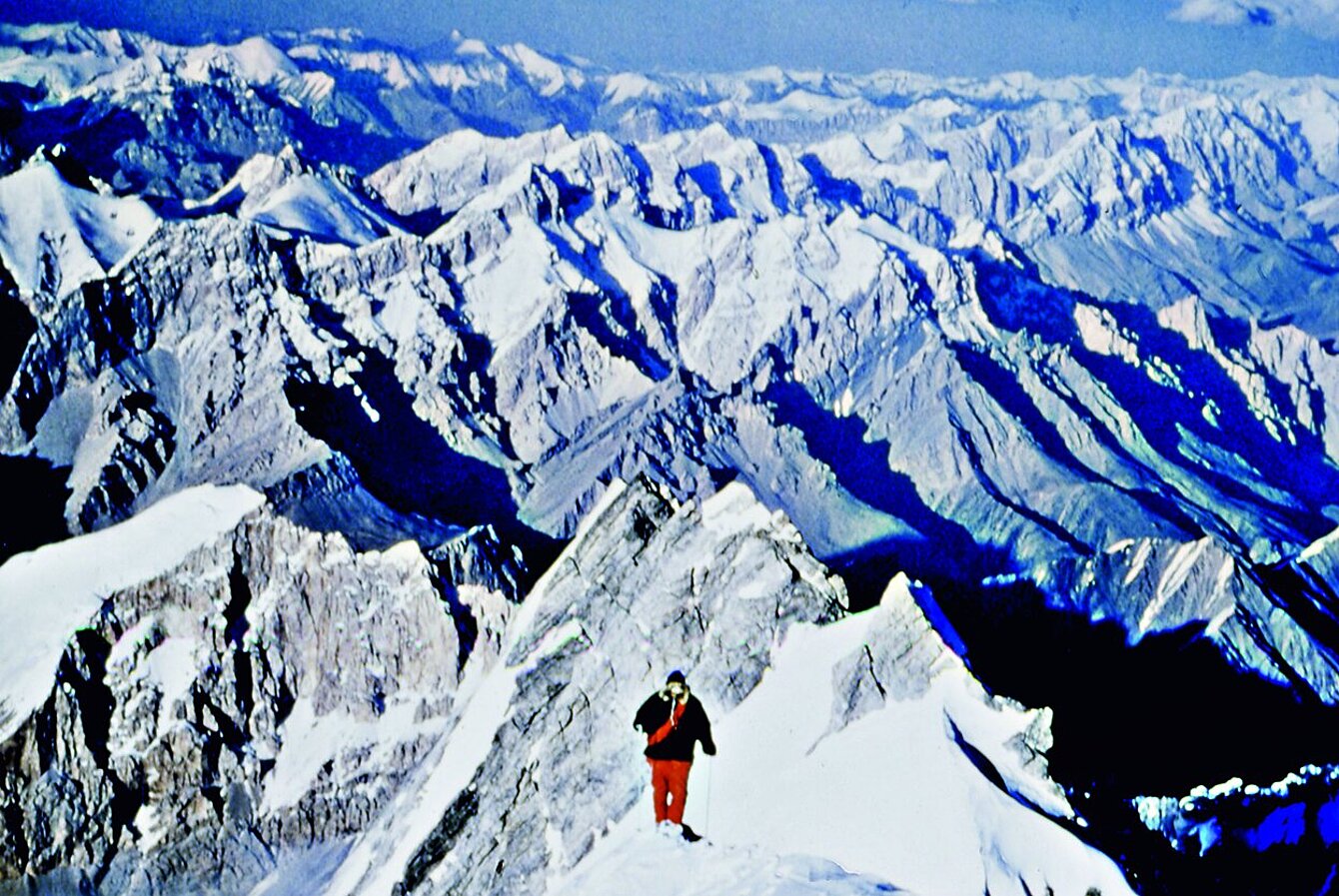Photograph of a man with red trousers and black jacket walking away from the camera. In the background white mountain peaks can be seen up to the horizon.