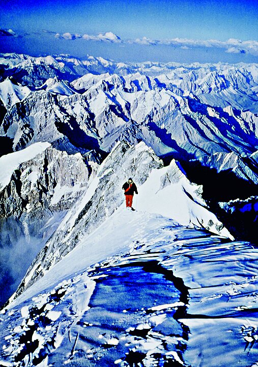 Fotografie von einem Mann mit roter Hose und schwarzer Jacke, der sich laufend von der Kamera entfernt. Im Hintergrund sind bis an den Horizont weiße Bergspitzen zu sehen.