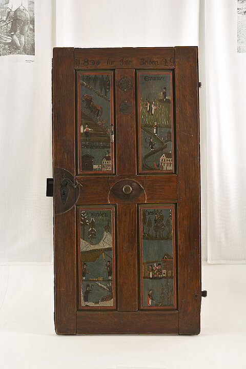 
A wooden door is displayed on a white base. The door is decorated with dark colors depicting scenes from the four seasons.