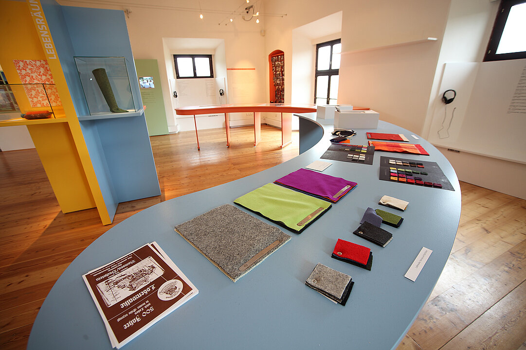 Ausstellungsansicht von einem Raum. In der Mitte steht ein bananenförmiger langer hellblauer Tisch, auf dem verschiedene farbige Stoffe aus Leinen liegen.