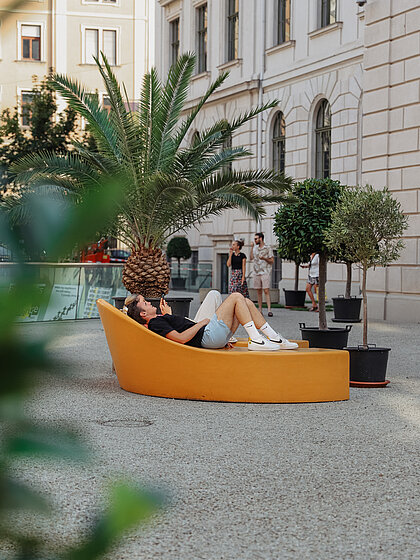 Two people are lying on the JOA at the Joanneumsviertelplatz in summer. Around them are palm trees and other plants.