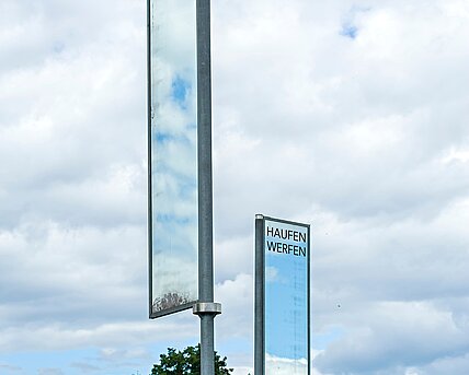 Auf einem Hügel zwischen zwei Wegen stehen zwei Spiegel auf Stangen. Durch ihre ständige Drehung im Wind geben sie stetig anderes wieder. Das sich in ihnen Spiegelnde symbolisiert einen Blick in zwei unterschiedliche Welten. Für die Momente, in denen sich die Spiegel ineinander spiegeln, entsteht eine Verbindung dieser beiden Welten.