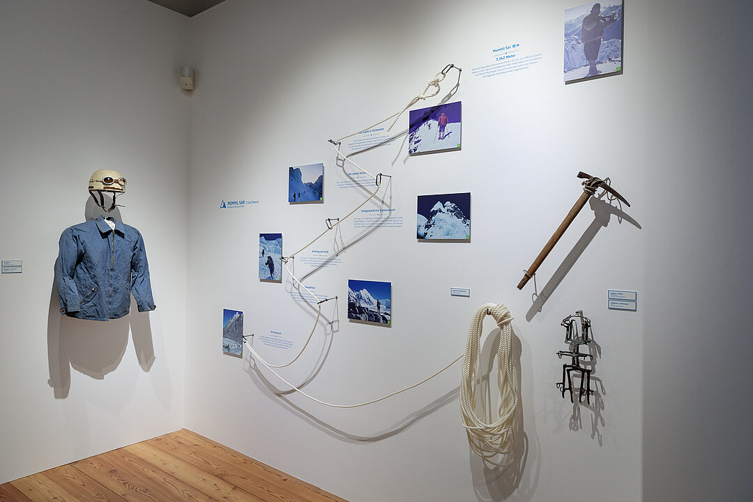 Ausstellungsansicht: An einer Wand hängen Fotografien, die mit einem Seil verbunden sind. Daneben hängt ein Outfit mit einer hellblauen Jacke und einem weißen Helm.