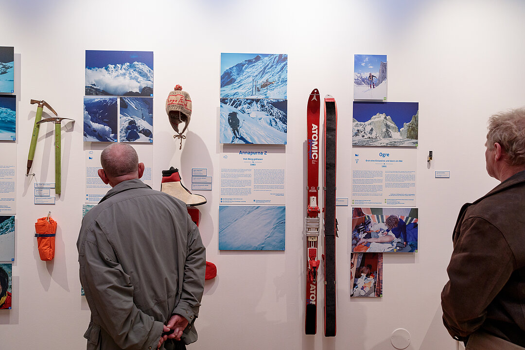 Foto von der Ausstellungseröffnung. Menschen stehen in einem Raum und betrachten Objekte (z.B. Schier) und Fotografien, die an der Wand hängen.