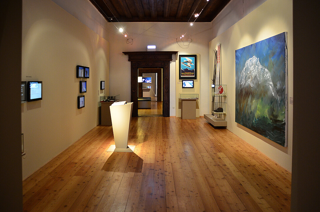 Ausstellungsansicht von einem Raum. Auf der rechten Seite hängt ein großes Gemälde, das abstrakt einen Berg darstellt. In der Mitte steht ein beleuchteter Sockel.