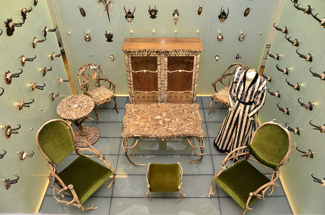 Exhibition view of a large display case with furniture made of antlers and horn. In the centre is a figurine of Countess Anna Lamberg. Antlers hang on the walls.