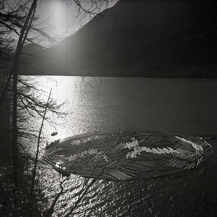 Alte schwarz-weiß Fotografie von einem See. Auf dem See schwimmen kreisförmig angeordnet Baumstämme.