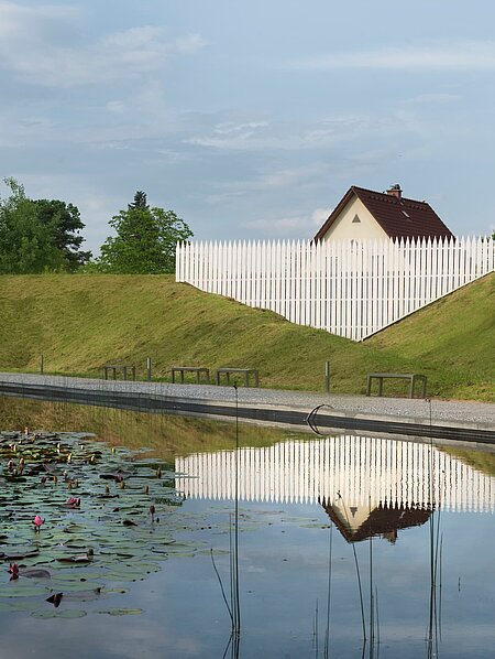 Ein übergroßer Gartenzaun, der die Umgebung außerhalb des Skulpturenparks miteinbezieht. Hinter dem Wall des Parks ragt das Dach eines typischen Einfamilienhauses hervor, das durch den übergroßen Zaun merkwürdig klein, beinahe versunken wirkt.