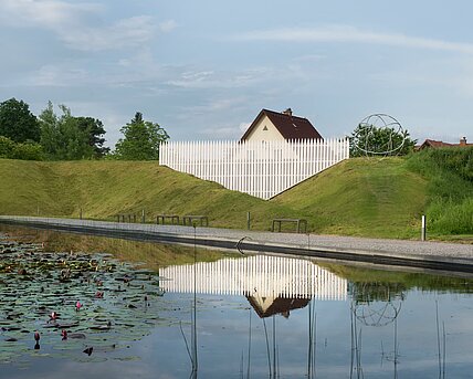 An oversized garden fence that incorporates the surroundings outside the sculpture park. The roof of a typical detached house protrudes from behind the wall of the park, appearing strangely small, almost sunken, due to the oversized fence.