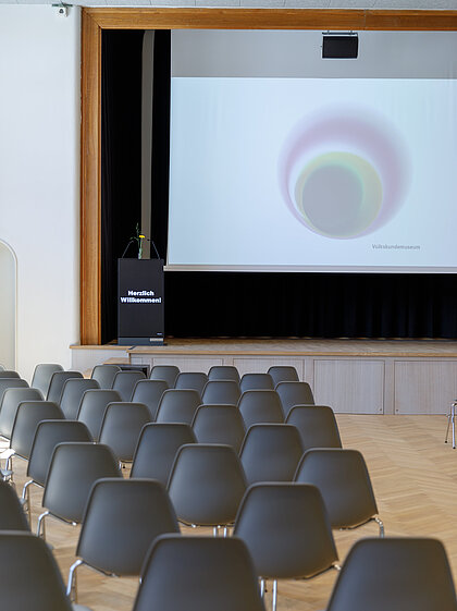 Chairs stand in the direction of the stage in the Heimatsaal, on it is a lectern as well as a screen.