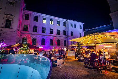 Belebter Adventmarkt am Abend im Hof eines historischen Gebäudes mit farbiger Fassadenbeleuchtung, geschmückten Weihnachtsbäumen, Marktständen, Lichterketten und Besucher*innen unter großen beleuchteten Schirmen.