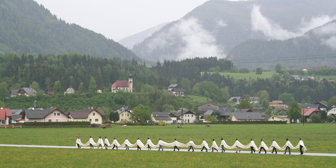 A group of men walk in a line through a foggy mountain landscape. All the men are tied with a white ribbon that lies over their shoulders.