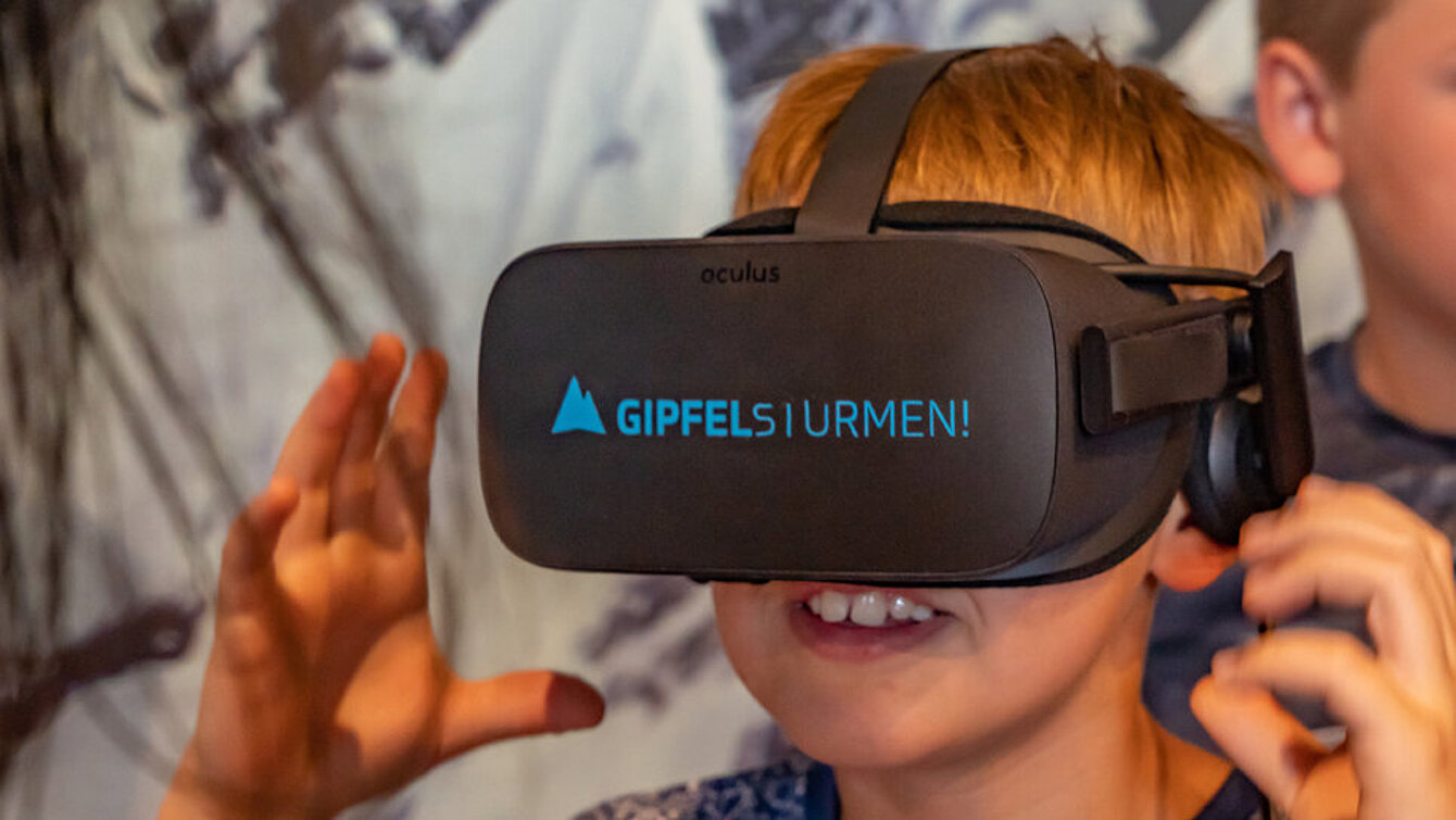 Ein Junge trägt eine schwarze VR-Brille mit der blauen Beschriftung Gipfelstürmen!