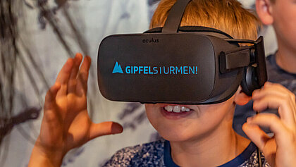 Ein Junge trägt eine schwarze VR-Brille mit der blauen Beschriftung Gipfelstürmen!