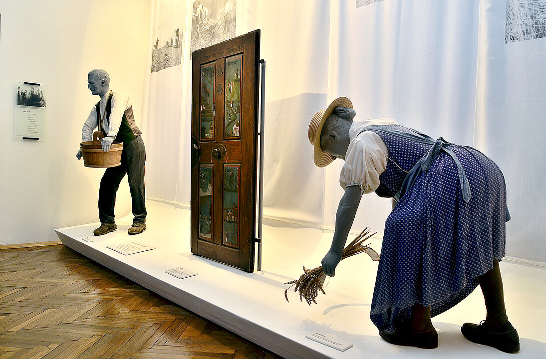 Photo of an exhibition room. On a long pedestal is a door. This is painted with motifs from the four seasons. To the right and left of the door are two mannequins in peasant clothes, working.