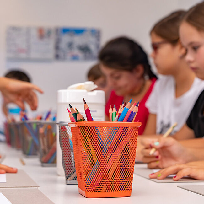 Kinder sitzen um einen Tisch. In der Mitte steht ein orangener Stiftebehälter mit bunten Stiften.