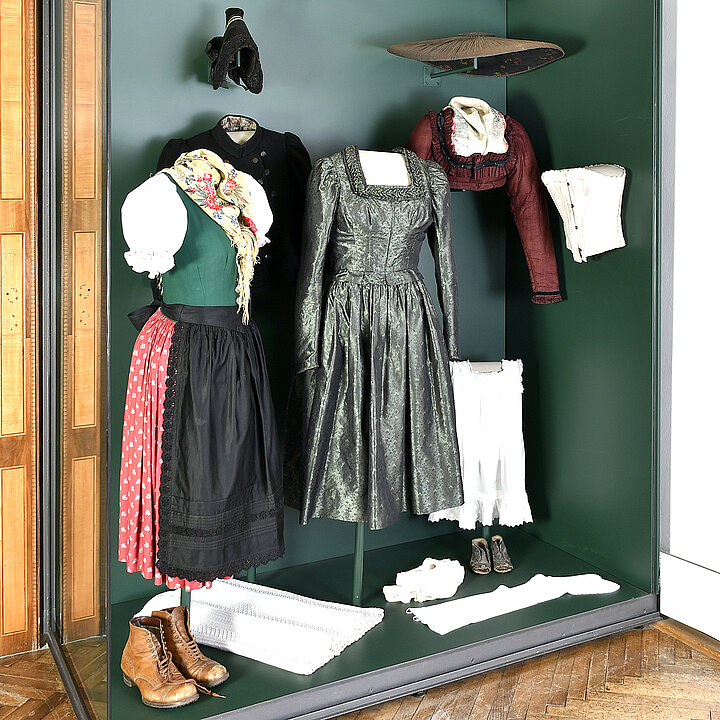 Eine Glasvitrine mit grünem Hintergrund. Darin sind Trachten zu sehen. Z.B. ein grünes Festtagskleid oder ein Dirndl mit schwarzer Schürze und rotem Rock. 