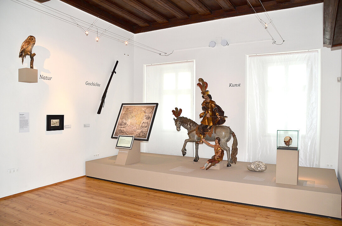 In einem Raum steht auf einem Sockel eine große Skulptur aus Holz. Sie zeigt die Figur des Heiligen Matins, der auf einem Pferd reitet unter ihm sitzt ein Mann, der bettelt.