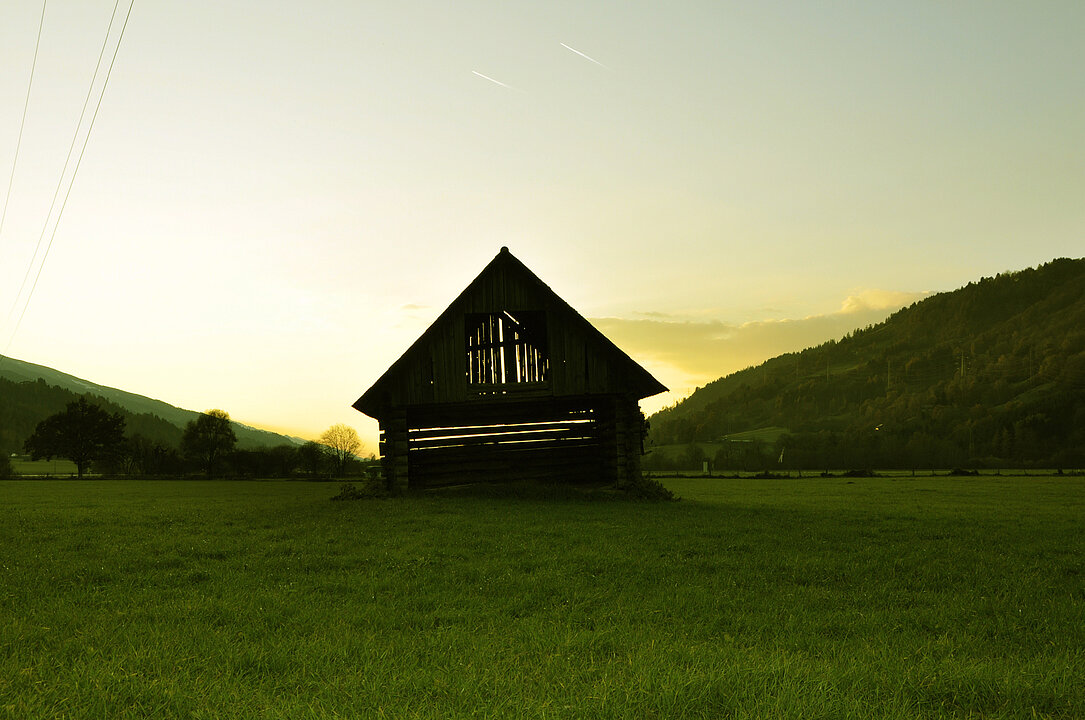 Fotografie von einem Heustadl. Im Hintergrund geht die Sonne zwischen den Hügeln auf.